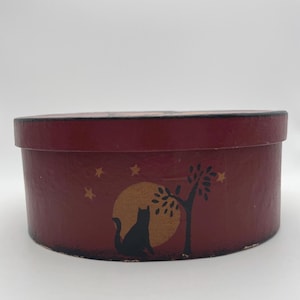 May include: A burgundy oval box with a black cat silhouette sitting in front of a gold moon. A tree and stars are also depicted on the box. The box is a decorative storage container.