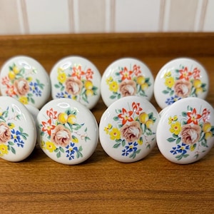 May include: A set of eight round white ceramic cabinet knobs, each featuring a colorful floral pattern with a central pink rose, yellow buds, small blue flowers, and green leaves arranged in a bouquet on the front surface of the hardware.