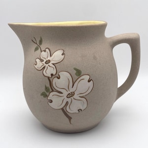 May include: A tan ceramic pitcher with a cream-colored floral design. The pitcher has a curved handle and a yellow rim. The floral design features white flowers with brown stems and green leaves. The pitcher is likely vintage.