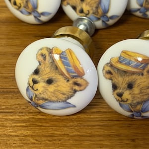 May include: A set of round white ceramic cabinet knobs featuring a printed illustration of a brown teddy bear wearing a yellow straw hat with blue stripes and a blue neckerchief. Each knob has a metallic gold-toned base and a smooth glossy finish.