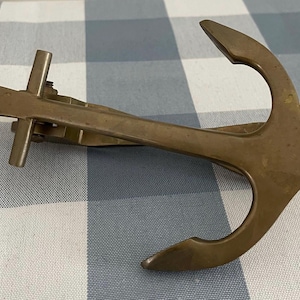 May include: A vintage brass anchor-shaped tie clip. The clip features a classic anchor design with a central bar and two arms. The brass has a warm, aged patina, suggesting its vintage origin. The clip is designed to secure a tie.