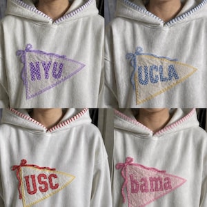 May include: White hooded sweatshirts with pennant patches. Each pennant displays a university name: NYU, UCLA, USC, and bama. The pennants are in various colors, with decorative stitching and bows.