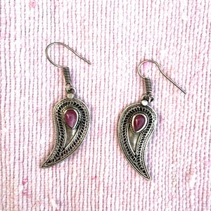 Puede incluir: Un par de pendientes plateados con diseño de cachemira. Cada pendiente presenta un marco ornamental en forma de lágrima con una pequeña piedra preciosa rosa ovalada en el centro. Cierre de gancho.