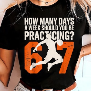 Op de afbeelding: Zwart t-shirt met de tekst "HOW MANY DAYS A WEEK SHOULD YOU BE PRACTICING?" in het wit. Daaronder de oranje cijfers "67" met een wit silhouet van een basketballer. Het shirt is een casual kledingstuk.