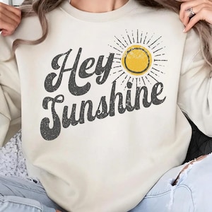 Puede incluir: Sudadera color crema con la frase "Hey Sunshine" en letras grises desgastadas. Un sol amarillo con rayos está encima de la palabra "Sunshine". La sudadera es de estilo casual y cómodo.
