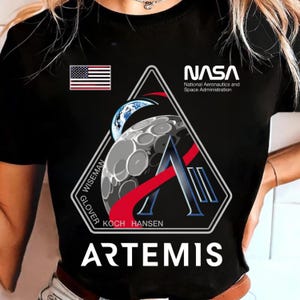 May include: Black t-shirt with the NASA logo and the Artemis mission patch. The patch shows the American flag, a stylized moon, and the crew names: Wiseman, Glover, Koch, and Hansen. The word "ARTEMIS" is printed in large white letters.