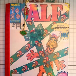 May include: A Marvel ALF comic book featuring a holiday-themed cover with the character wrapped in green ribbon. A gold-toned metal bookmark with a small red gift charm and a green ornament charm is attached to the top of the comic book.