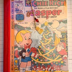 May include: A Richie Rich and Casper comic book with a red spine and gold corner protectors. A gold-toned metal bookmark chain is clipped to the top, featuring a star charm, a small red bulb, and a gold-toned ghost-shaped charm.