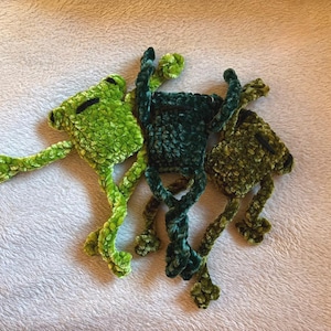Könnte beinhalten: Drei handgefertigte Häkelfrosch-Spielzeuge in Grüntönen. Die Frösche haben ausgestreckte Arme und Beine mit schwarzen Augen. Die Plüschtiere sind aus einem weichen, strukturierten Garn gefertigt und liegen auf einer weißen Oberfläche.