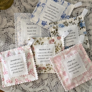 May include: A collection of square fabric sachets featuring floral patterns in blue, pink, and brown. Each sachet has a white center patch with printed Bible verses including Proverbs 3:5, Psalm 46:10, Psalm 46:1, Matthew 11:28, Jeremiah 29:13, and 1 Peter 5:7.