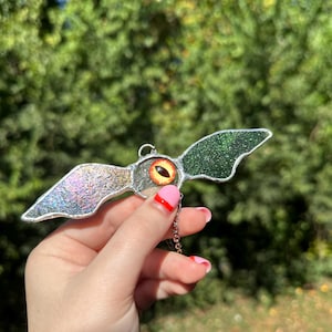 May include: A stained glass bat-shaped ornament featuring an eye design. The wings are iridescent and green, with a silver metal border. A small chain is attached for hanging. The background is blurred green foliage.