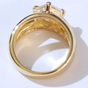May include: A gold-colored ring with a large, clear, rectangular-cut gemstone. The ring has a thick band and a detailed setting that holds the gemstone securely. The ring is shown against a white background.