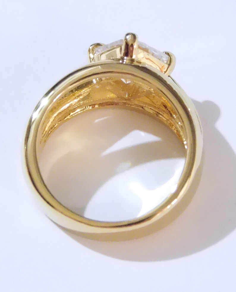 May include: A gold-colored ring with a large, clear, rectangular-cut gemstone. The ring has a thick band and a detailed setting that holds the gemstone securely. The ring is shown against a white background.