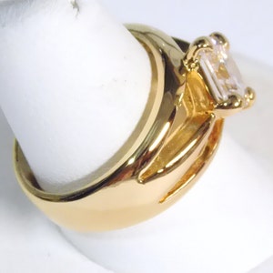 May include: A gold-colored ring with a large, rectangular, clear gemstone. The ring has a thick band and a detailed setting around the gemstone. The ring is displayed on a white surface.