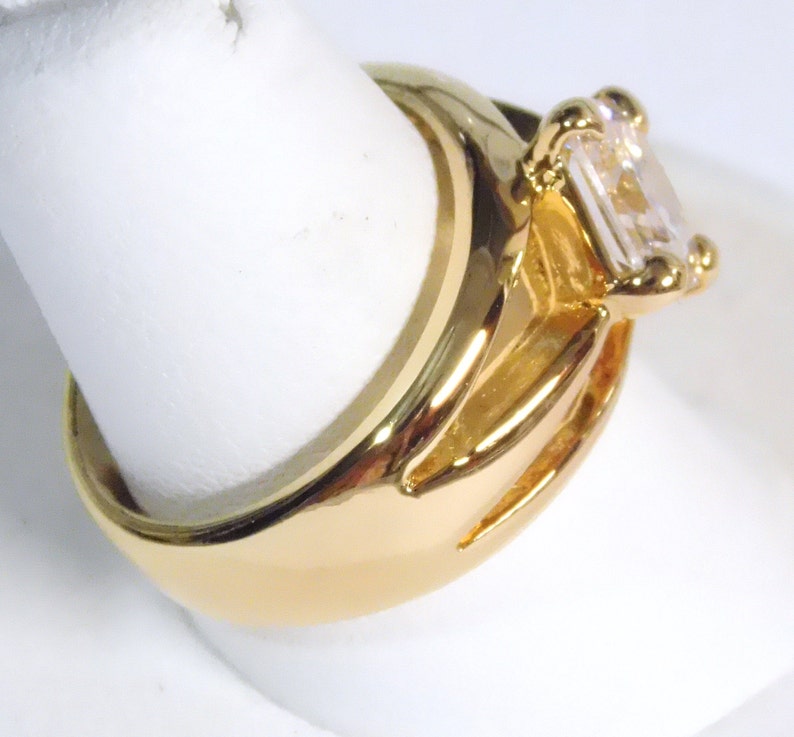 May include: A gold-colored ring with a large, rectangular, clear gemstone. The ring has a thick band and a detailed setting around the gemstone. The ring is displayed on a white surface.