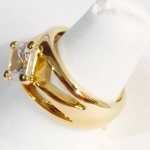 May include: A gold-colored ring with a large, square-cut, clear gemstone. The ring has a split shank design with three curved prongs on each side, and small gold accents around the gemstone. The ring is displayed on a white surface.