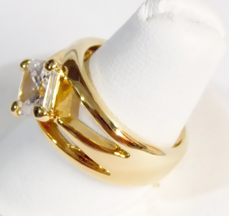 May include: A gold-colored ring with a large, square-cut, clear gemstone. The ring has a split shank design with three curved prongs on each side, and small gold accents around the gemstone. The ring is displayed on a white surface.