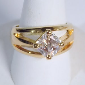 May include: A gold-toned ring featuring a large, square-cut, clear gemstone. The ring has a wide band with a split design and small gold accents around the gemstone. The ring is displayed on a white stand.
