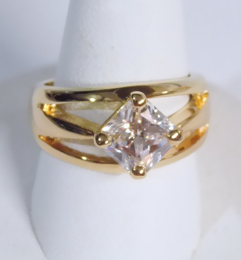 May include: A gold-toned ring featuring a large, square-cut, clear gemstone. The ring has a wide band with a split design and small gold accents around the gemstone. The ring is displayed on a white stand.