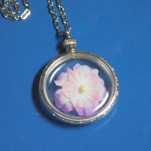 May include: A silver-toned necklace with a round pendant. The pendant encases a delicate flower with white, pink, and purple petals and a yellow center. The chain is a fine link style, and the pendant has a clear, glass-like cover.