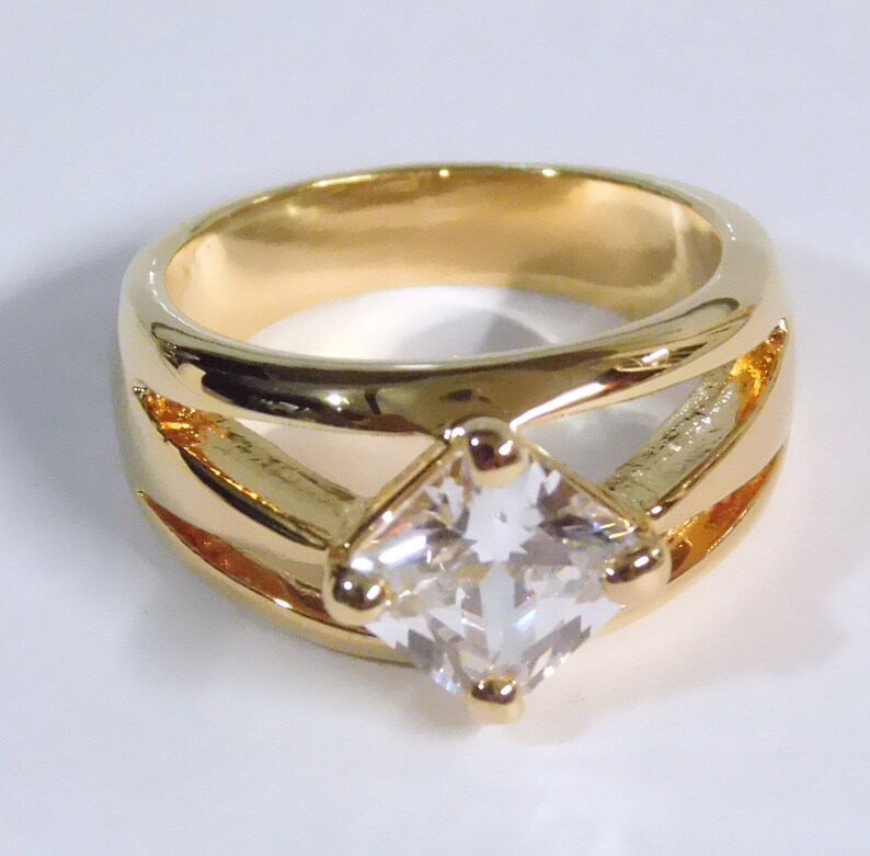 May include: A gold-colored ring with a large, square, clear gemstone at the center. The ring has a thick band with cut-out design elements on the sides. The gemstone is held in place by four small prongs. The ring is likely made of gold or gold-plated metal.