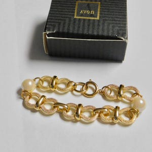 May include: Gold-tone bracelet with textured links and faux pearls. The bracelet is next to a black box with gold lettering that reads "AVON". The bracelet has a clasp closure.