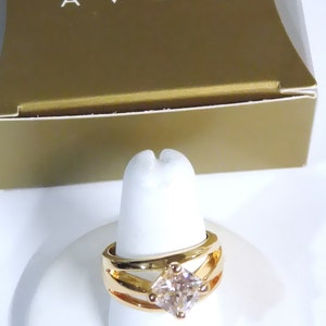 May include: Gold-toned ring with a square, light-colored gemstone. The ring has a split-shank design and is displayed on a white ring holder. The word "AVON" is visible on the gold-colored box in the background.