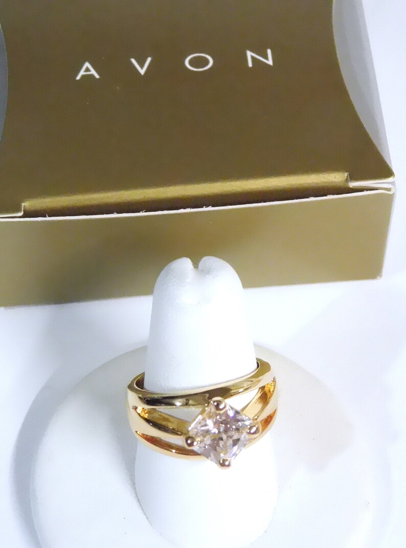 May include: Gold-toned ring with a square, light-colored gemstone. The ring has a split-shank design and is displayed on a white ring holder. The word "AVON" is visible on the gold-colored box in the background.