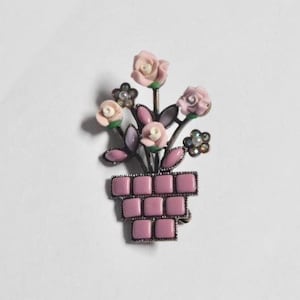 May include: A vintage brooch featuring a pink flower arrangement in a square pot. The brooch has a silver-tone metal frame, with pink roses, leaves, and small pearl-like accents. The pot is made of pink square tiles.