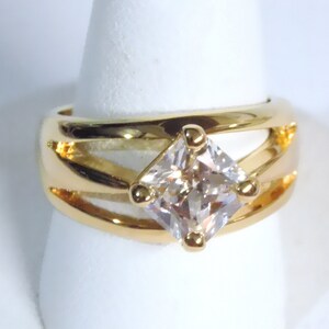May include: A gold-toned ring featuring a large, square, clear gemstone at the center. The ring has a split-shank design with a polished finish. The gemstone is held in place by four small gold prongs. The ring is displayed on a white surface.