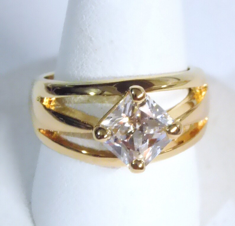 May include: A gold-toned ring featuring a large, square, clear gemstone at the center. The ring has a split-shank design with a polished finish. The gemstone is held in place by four small gold prongs. The ring is displayed on a white surface.