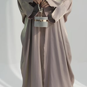 May include: A flowing, taupe-coloured abaya with wide sleeves and a matching head covering. The abaya is paired with black sandals and a small, rectangular, glittery clutch with a pearl handle. The outfit is elegant and modest.