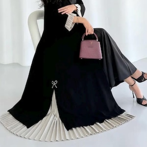 May include: A long-sleeved black dress featuring a white pleated hem and matching white pleated cuffs. The dress includes a small white bow detail on the side slit and is paired with a structured mauve handbag and black strappy high-heeled sandals.