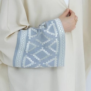 May include: Cream-coloured abaya with wide sleeves featuring a light blue cuff. The cuff is decorated with a geometric pattern in silver and white. The abaya is a loose-fitting garment, often worn in the Middle East and North Africa.