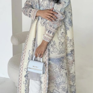 Può includere: Un lungo cappotto plissettato color crema con un disegno a mappa in blu e grigio. Il cappotto è indossato con pantaloni bianchi e una borsetta azzurra. La borsetta ha un manico superiore e una chiusura color oro. Una piccola fotocamera è visibile sullo sfondo.