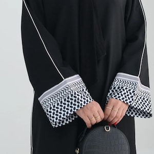Könnte beinhalten: Schwarze Abaya mit weißem Besatz an Ärmeln und Seiten. Die Manschetten haben ein schwarz-weißes geometrisches Muster. Eine schwarze, runde Handtasche mit Krokodiltextur wird gehalten.