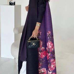 May include: A black pleated long coat with a purple floral design on the back. The coat is paired with white trousers and clear heels. A small black handbag with a gold chain is held in the hand.