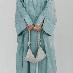 May include: A long, flowing, light teal abaya with wide sleeves and a matching head covering. The abaya is paired with a white top and trousers. A silver, beaded handbag is held in front.