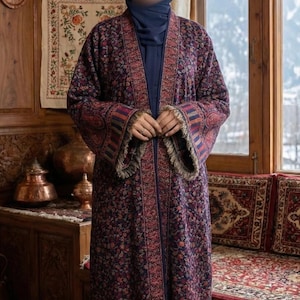 May include: A long, open-front coat featuring a dark purple base with an intricate, multi-coloured floral and paisley pattern. The garment has long sleeves with striped cuffs and a fringed hem, worn over a solid dark blue shirt and head covering.