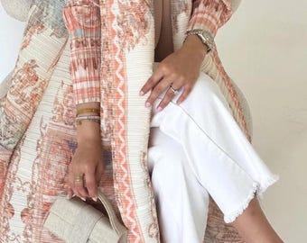 Könnte beinhalten: Ein langer, plissierter Mantel mit einem Muster in Pfirsich, Creme und Blau. Der Mantel wird mit einer weißen Hose und einer kleinen, hellfarbigen Handtasche getragen. Das Outfit wird durch transparente Absatzsandalen ergänzt. Das Muster des Mantels ist detailliert.