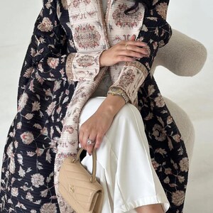 K&ouml;nnte beinhalten: Eine lange, schwarze Jacke mit Blumenmuster und cremefarbenem Zierrat und Futter. Die Jacke wird mit einer wei&szlig;en Hose und einer kleinen, hellbraunen Handtasche kombiniert. Das Blumenmuster der Jacke umfasst rosa und cremefarbene Blumen.