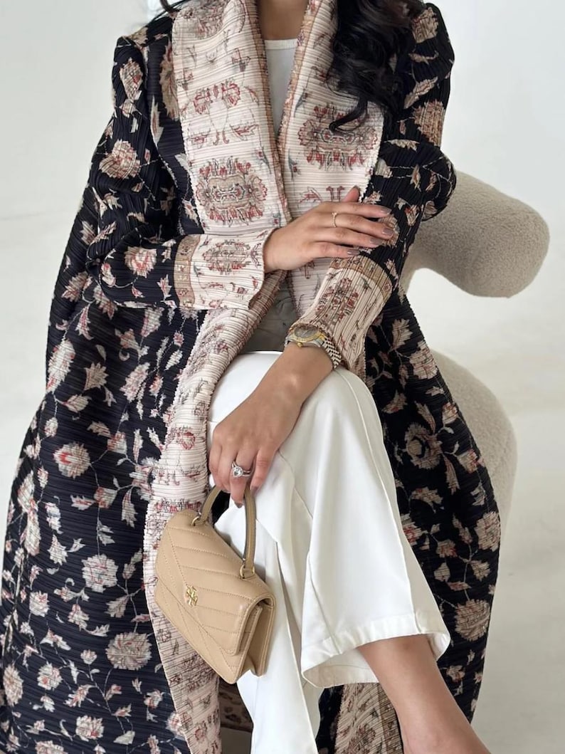 K&ouml;nnte beinhalten: Eine lange, schwarze Jacke mit Blumenmuster und cremefarbenem Zierrat und Futter. Die Jacke wird mit einer wei&szlig;en Hose und einer kleinen, hellbraunen Handtasche kombiniert. Das Blumenmuster der Jacke umfasst rosa und cremefarbene Blumen.