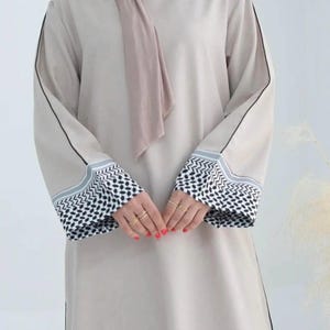 May include: A beige abaya featuring long sleeves with black and white patterned cuffs, a thin black line running down the length of each sleeve, and a matching beige hijab draped around the neck, creating a cohesive and neutral-toned ensemble.
