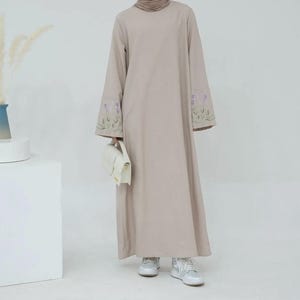 May include: A floor-length beige abaya dress featuring long sleeves with embroidered lavender floral details at the cuffs. The garment has a high neckline and a loose, flowing silhouette. A small white structured handbag is held at the side.