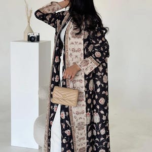 K&ouml;nnte beinhalten: Langer, schwarzer Mantel mit floralem Muster und beigefarbenem und floralem Besatz. Der Mantel wird &uuml;ber einem wei&szlig;en Outfit getragen und mit einer beigefarbenen Handtasche kombiniert. Eine kleine Kamera und eine Vase sind im Hintergrund zu sehen.