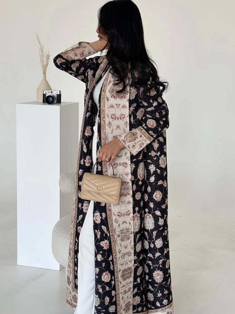 K&ouml;nnte beinhalten: Langer, schwarzer Mantel mit floralem Muster und beigefarbenem und floralem Besatz. Der Mantel wird &uuml;ber einem wei&szlig;en Outfit getragen und mit einer beigefarbenen Handtasche kombiniert. Eine kleine Kamera und eine Vase sind im Hintergrund zu sehen.