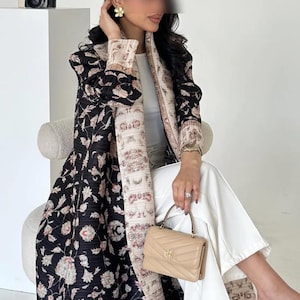K&ouml;nnte beinhalten: Ein langer, schwarzer Mantel mit floralem Muster und cremefarbenem, gemustertem Besatz. Der Mantel wird &uuml;ber einem wei&szlig;en Oberteil und einer Hose getragen. Eine kleine beige Handtasche wird in der Hand gehalten. Eine Vintage-Kamera steht auf einer wei&szlig;en Oberfl&auml;che.