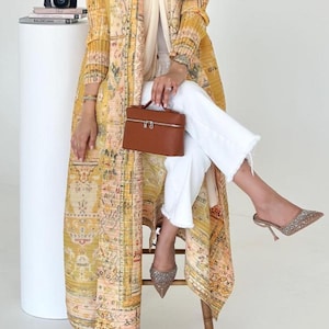 May include: A long, yellow patterned duster coat with a matching scarf, worn over a white top and pants. A brown handbag and embellished heels complete the look. A camera and books are on a white pedestal.