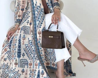 May include: A long, patterned coat in cream, blue, and brown tones. The coat features a detailed botanical design. A brown handbag with a gold clasp and sparkling high-heeled shoes complete the look. White pants are also visible.