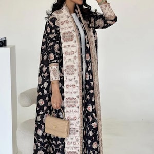 K&ouml;nnte beinhalten: Ein langer, schwarzer Mantel mit floralem Muster und einem cremefarbenen, gemusterten Rand und Futter. Der Mantel wird &uuml;ber einem wei&szlig;en Outfit getragen und mit einer kleinen, beigen Handtasche kombiniert. Das Blumenmuster enth&auml;lt rosa und cremefarbene Blumen.
